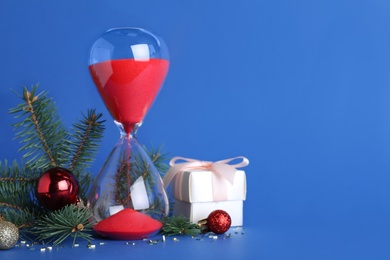 Photo of Hourglass, gift, fir tree branches with decor on blue background, space for text. Christmas countdown