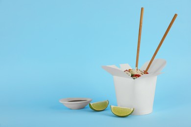 Photo of Box of vegetarian wok noodles with chopsticks, lime and soy sauce on light blue background. Space for text