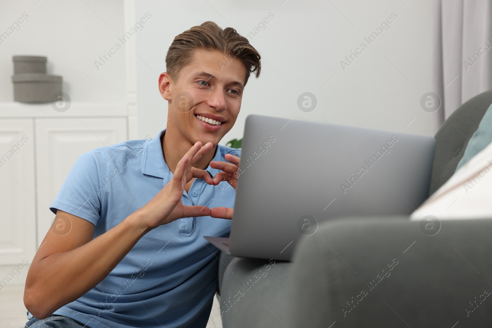 Photo of Happy young man having video chat via laptop and making heart with hands on sofa indoors. Long-distance relationship