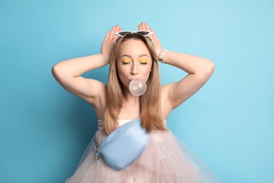 Fashionable young woman with bright makeup blowing bubblegum on light blue background