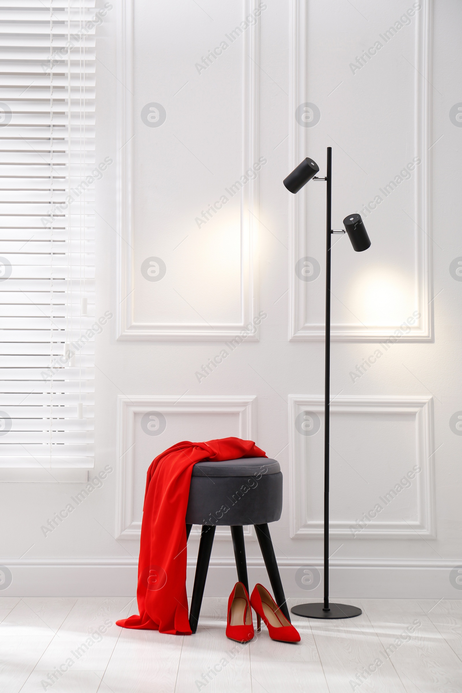 Photo of Stylish ottoman with dress and floor lamp near white wall in room. Interior design