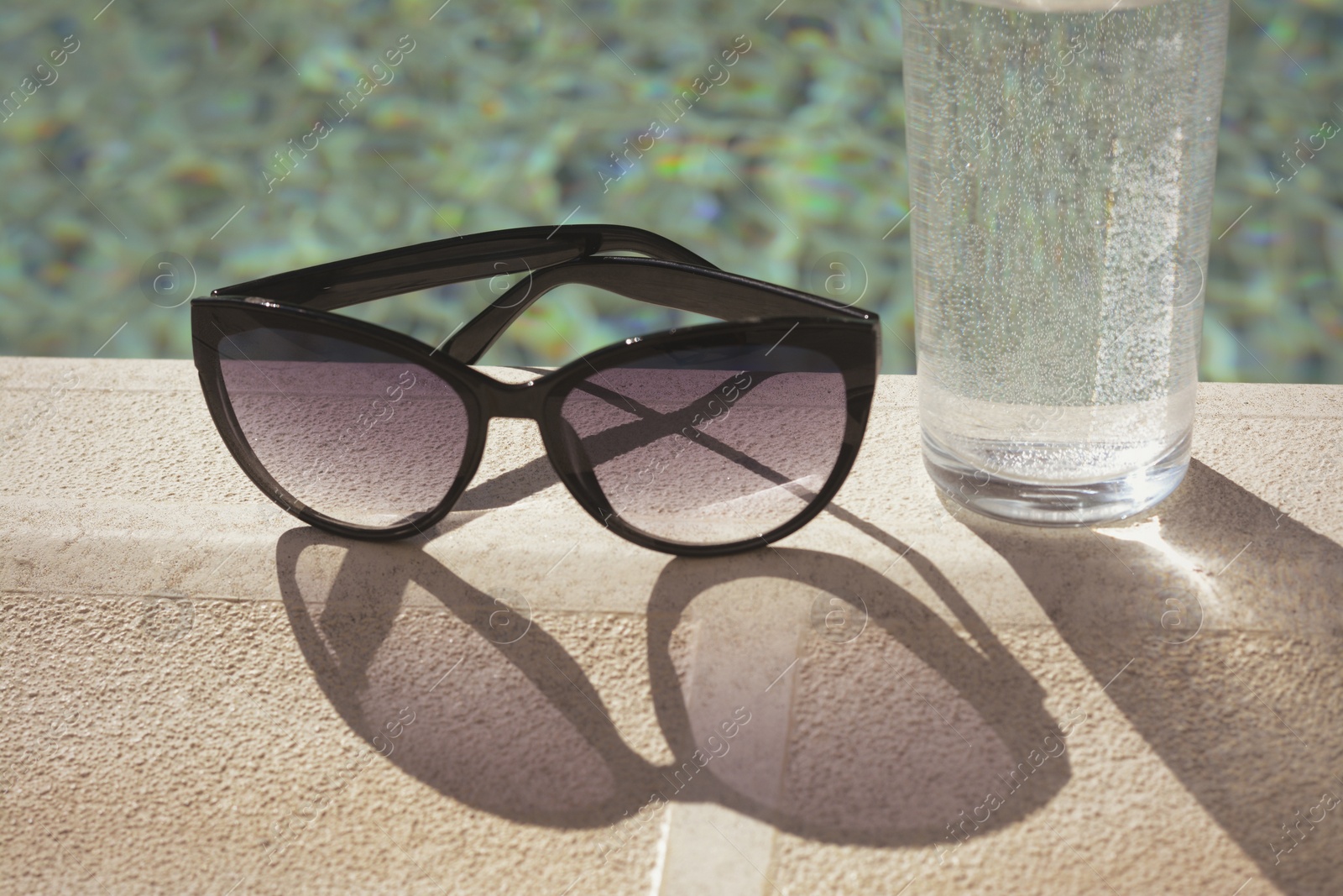 Photo of Stylish sunglasses and glass of water near outdoor swimming pool on sunny day, closeup