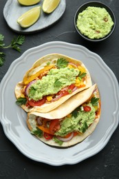 Photo of Delicious tacos with guacamole and vegetables served on black table, flat lay