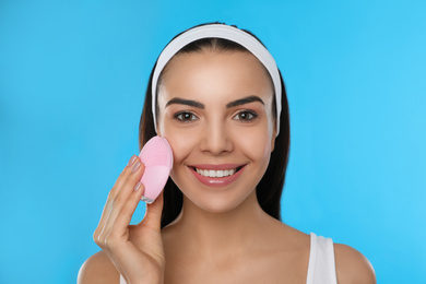 Photo of Young woman using facial cleansing brush on light blue background. Washing accessory