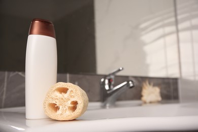 Natural loofah sponge and bottle of shower gel on washbasin in bathroom, space for text