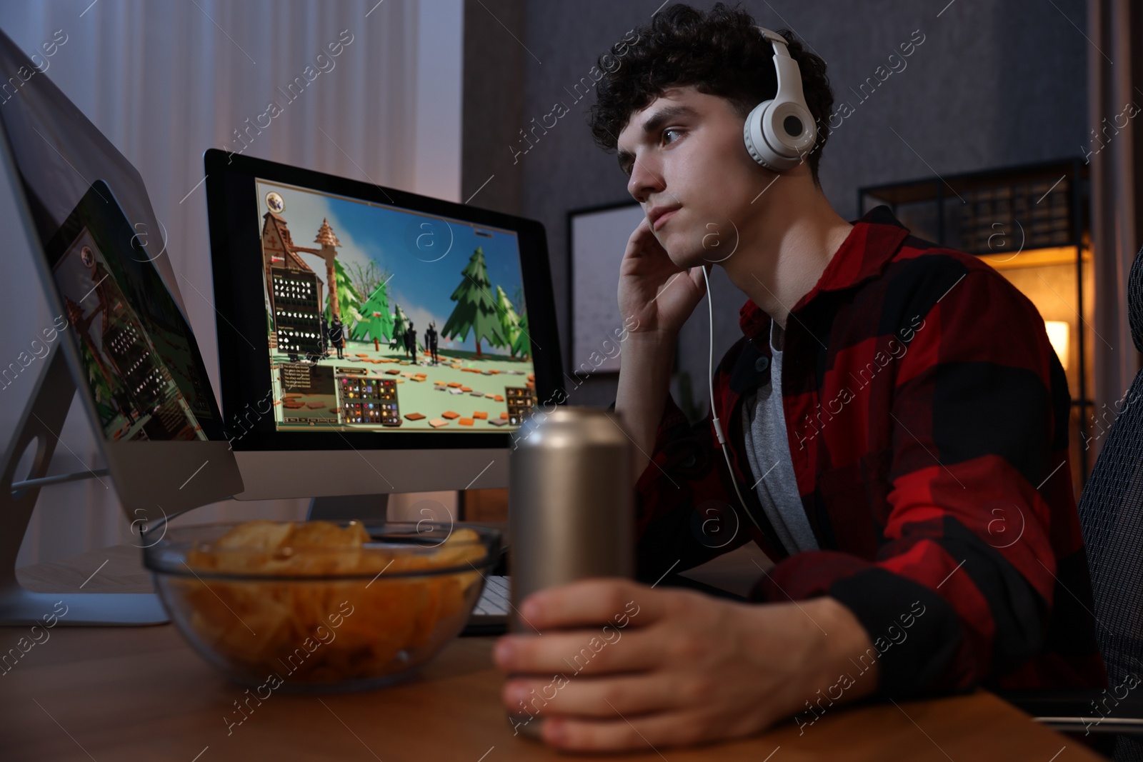 Photo of Young man with energy drink and headphones playing video game at wooden desk indoors