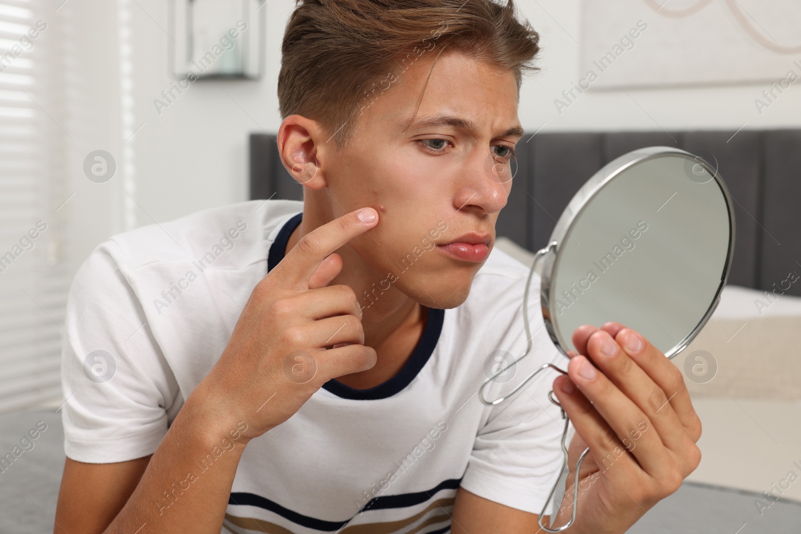 Photo of Upset young man looking at mirror and touching pimple on his face indoors. Acne problem