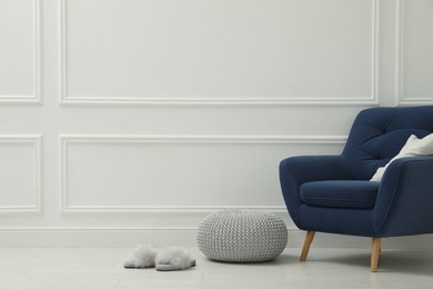 Knitted pouf, fuzzy slippers and armchair near white wall indoors. Space for text