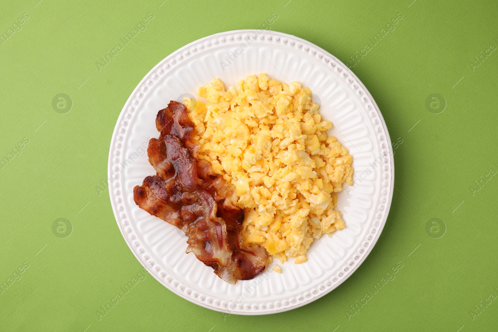 Photo of Delicious scrambled eggs with bacon in plate on light green background, top view