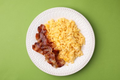 Delicious scrambled eggs with bacon in plate on light green background, top view