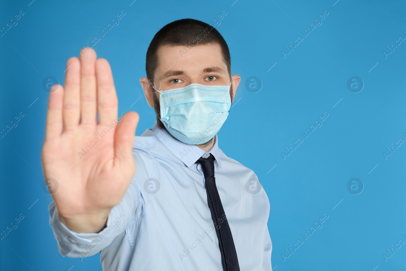 Photo of Man in protective mask showing stop gesture on light blue background, space for text. Prevent spreading of coronavirus