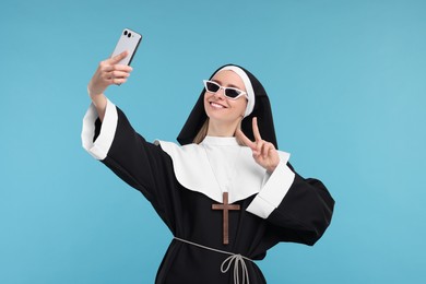 Photo of Happy woman in nun habit taking selfie and showing peace sign against light blue background