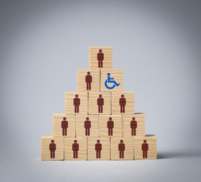 Image of Inclusive workplace culture. Pyramid of wooden cubes with human icons and one with international symbol of access on grey background