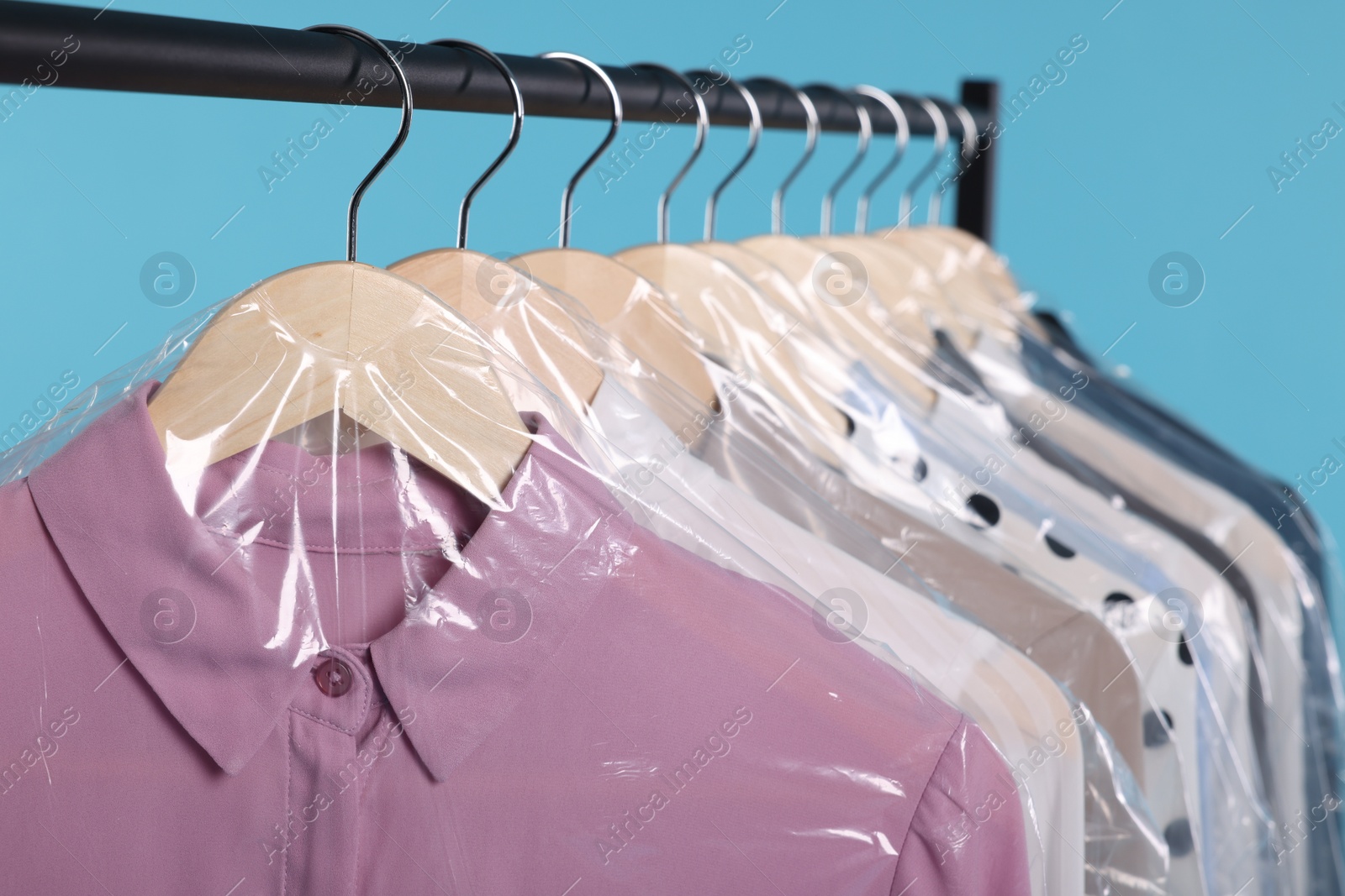 Photo of Dry-cleaning service. Many different clothes in plastic bags hanging on rack against light blue background, closeup