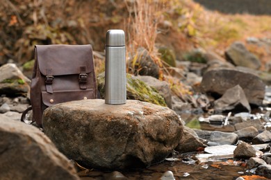 Metallic thermos and leather backpack on stone near river, space for text