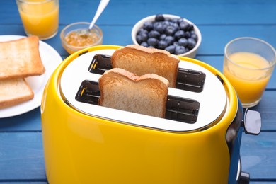 Photo of Yellow toaster with roasted bread, glasses of juice, blueberries and jam on blue wooden table, closeup