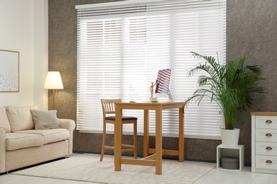 Photo of Comfortable workplace near window with blinds in room