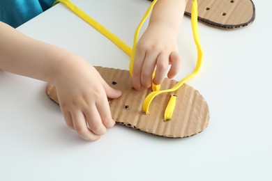 Little boy tying shoe lace using training cardboard template at white table, closeup