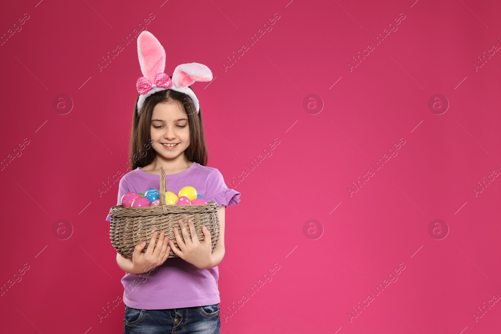 Photo of Little girl in bunny ears headband holding basket with Easter eggs on color background, space for text