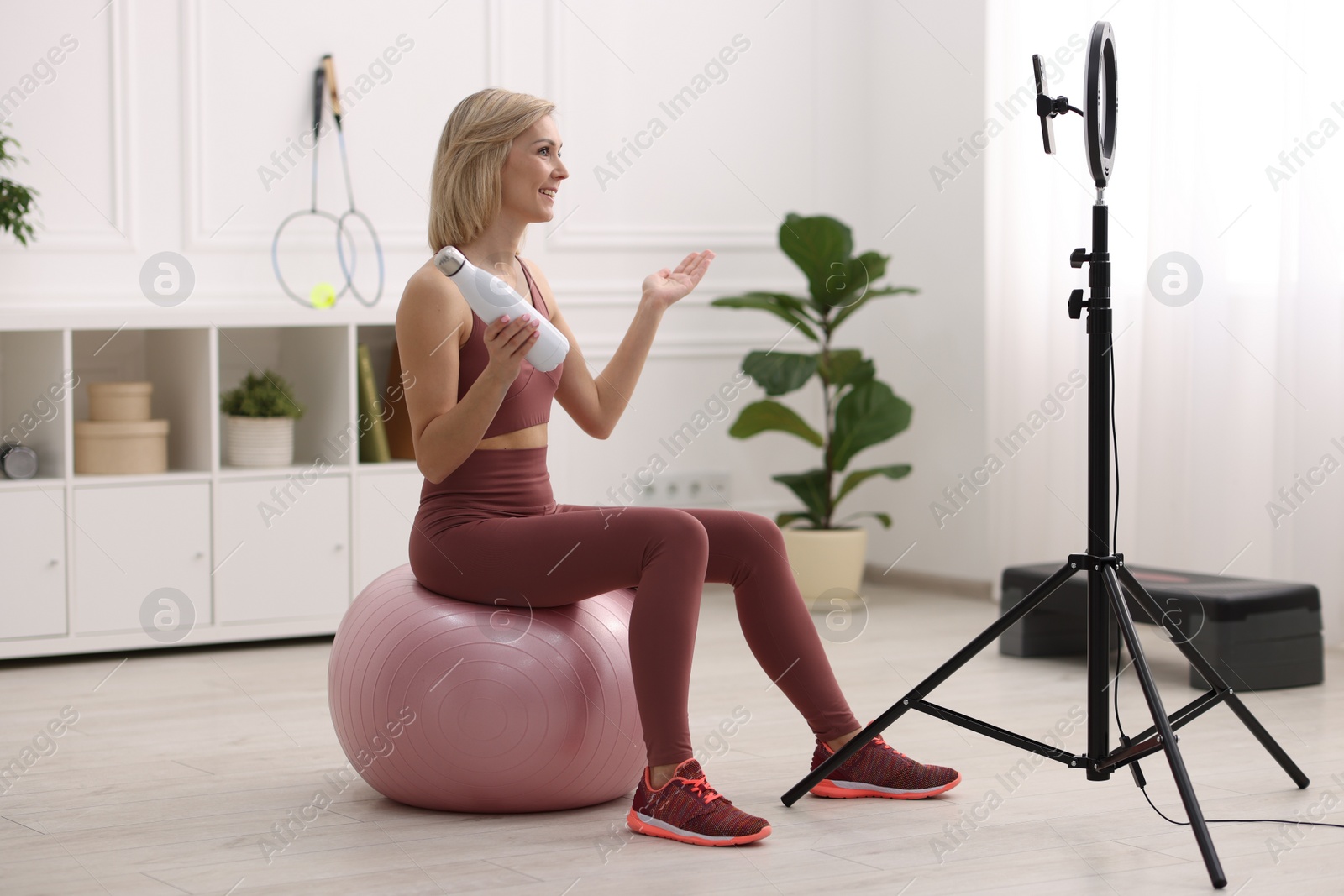 Photo of Smiling sports blogger holding bottle while streaming online fitness lesson at home