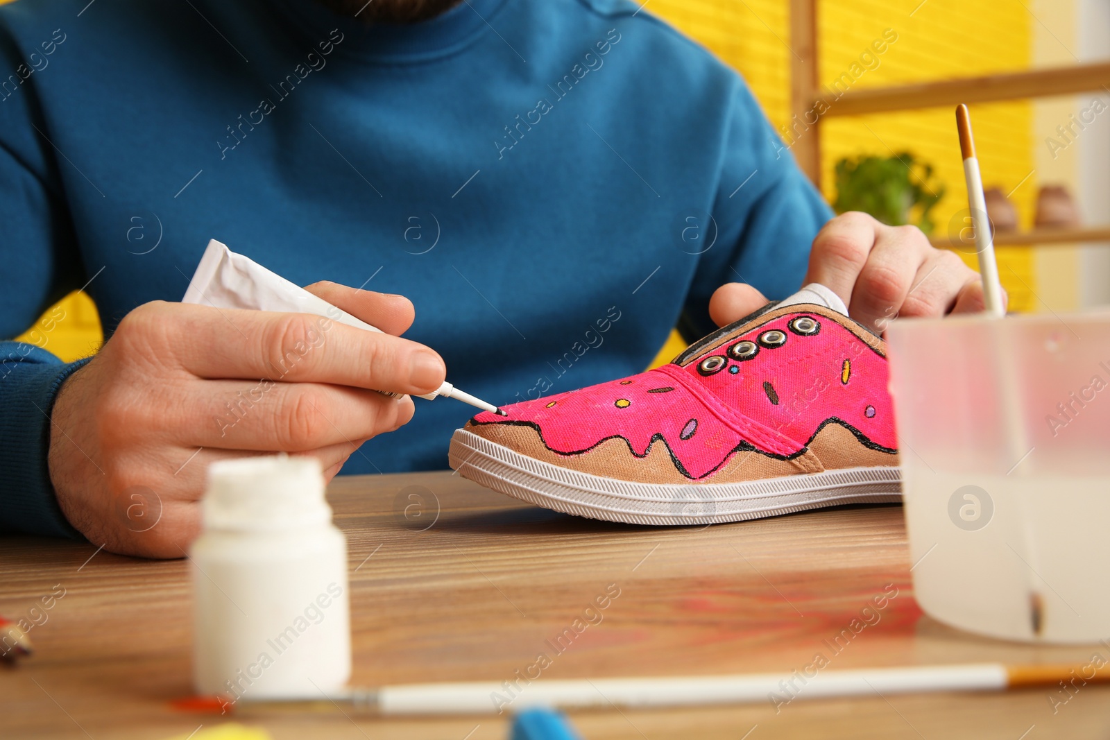 Photo of Man painting on sneaker at wooden table indoors, closeup. Customized shoes