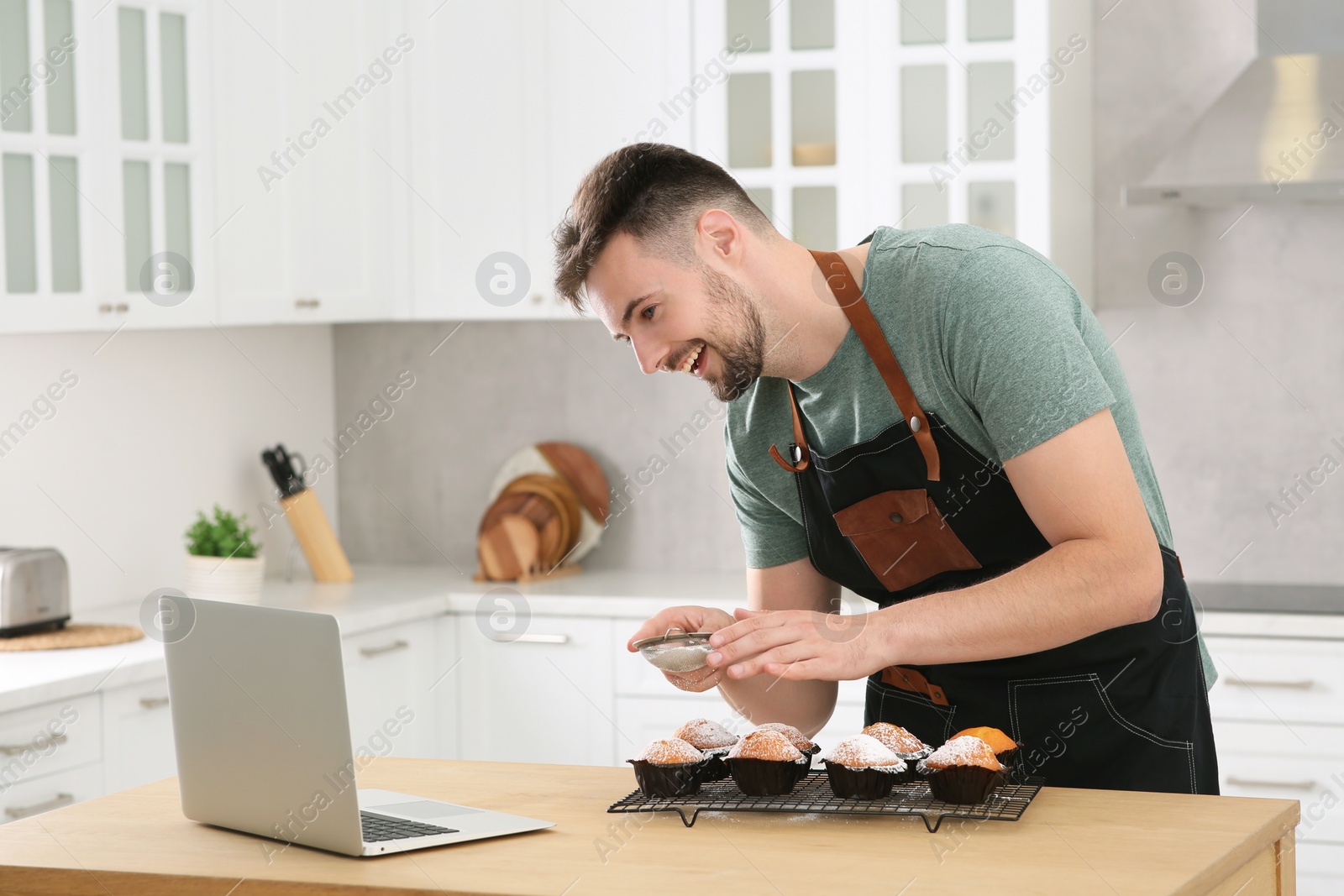 Photo of Man decorating muffins with powdered sugar while watching online cooking course via laptop in kitchen. Time for hobby