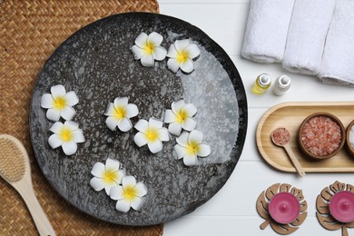 Bowl of water with flowers and different spa supplies on white wooden table, flat lay