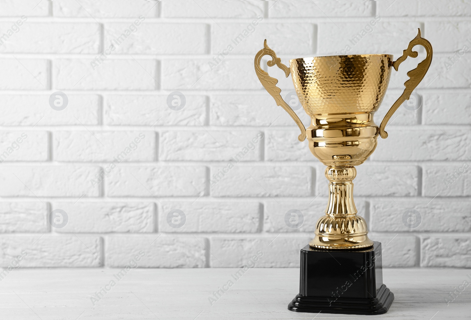 Photo of Golden trophy cup on table near white brick wall. Space for text