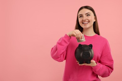 Photo of Happy woman putting dollar banknote into piggy bank on pink background, space for text