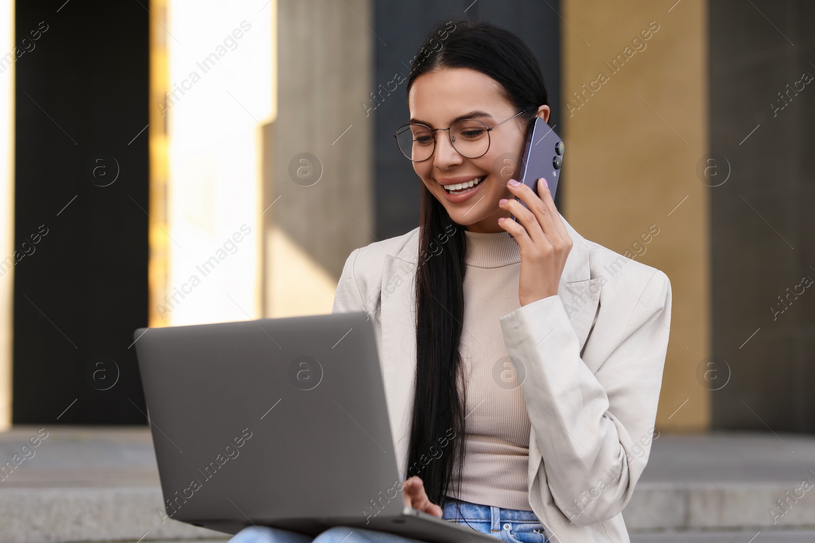 Photo of Happy young woman using modern laptop and talking on smartphone outdoors