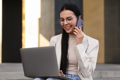 Photo of Happy young woman using modern laptop and talking on smartphone outdoors