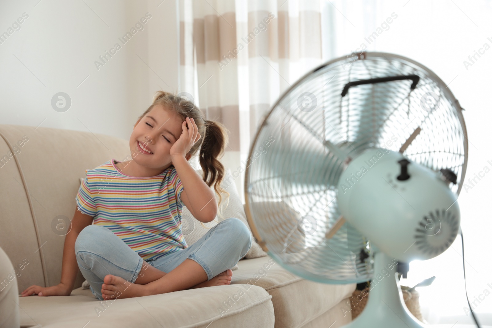 Photo of Little girl enjoying air flow from fan on sofa in living room. Summer heat