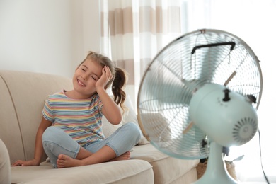 Photo of Little girl enjoying air flow from fan on sofa in living room. Summer heat