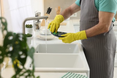 Man in protective gloves washing plate above sink in kitchen, closeup