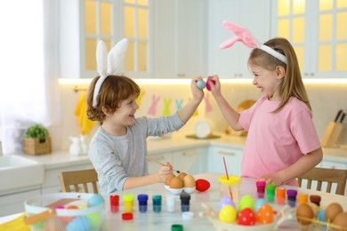 Photo of Easter celebration. Cute children with bunny ears having fun while painting eggs at white marble table in kitchen