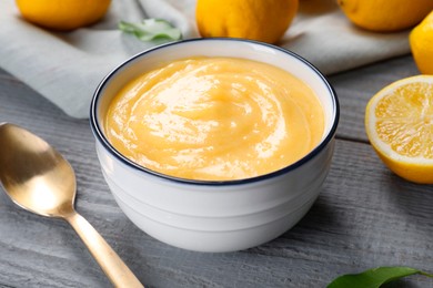 Delicious lemon curd in bowl, fresh citrus fruits and spoon on grey wooden table, closeup