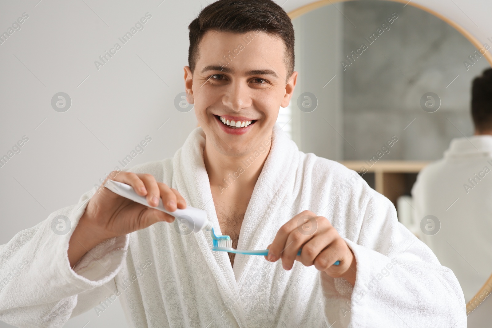 Photo of Happy man squeezing toothpaste from tube onto toothbrush in bathroom