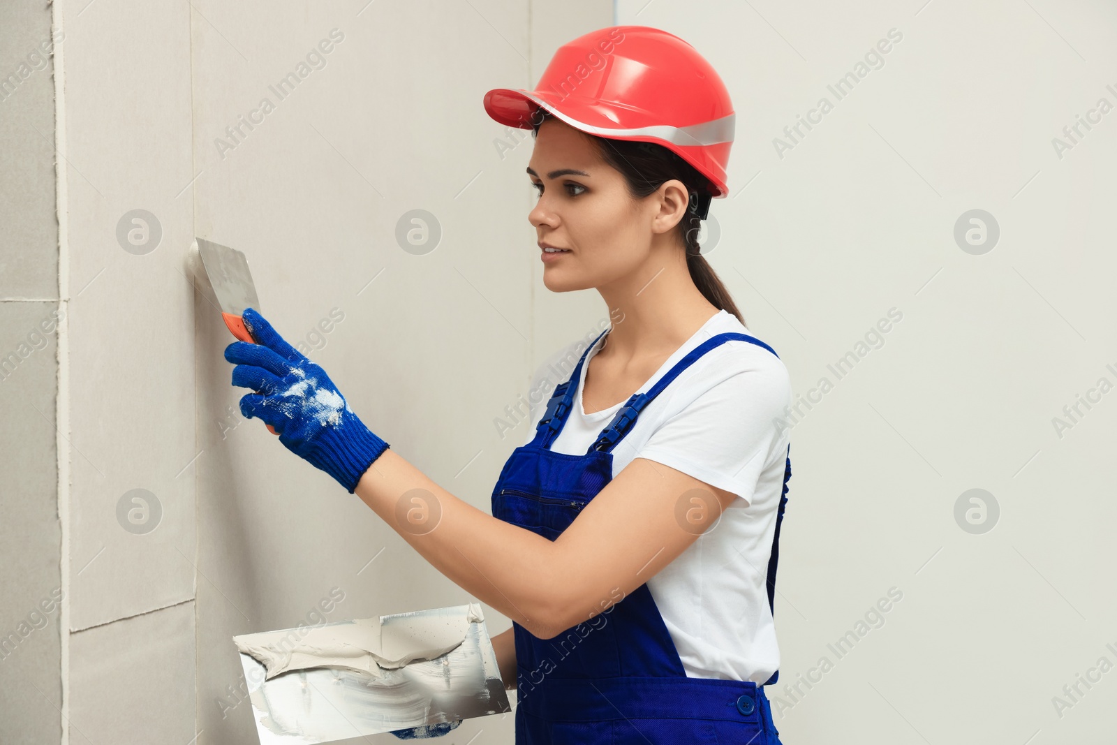 Photo of Professional worker in hard hat plastering wall with putty knives indoors