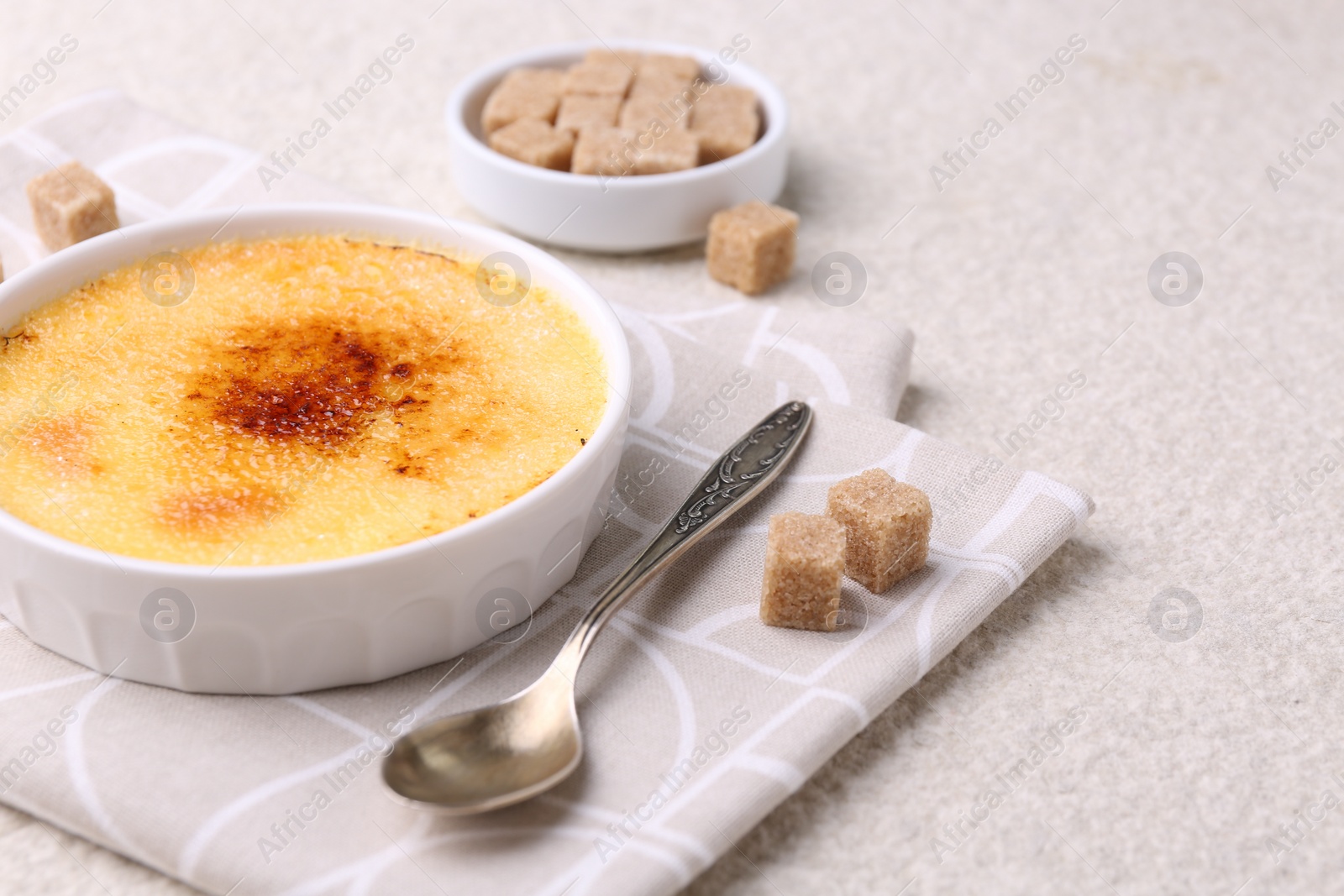 Photo of Delicious creme brulee in bowl, sugar cubes and spoon on light textured table, closeup