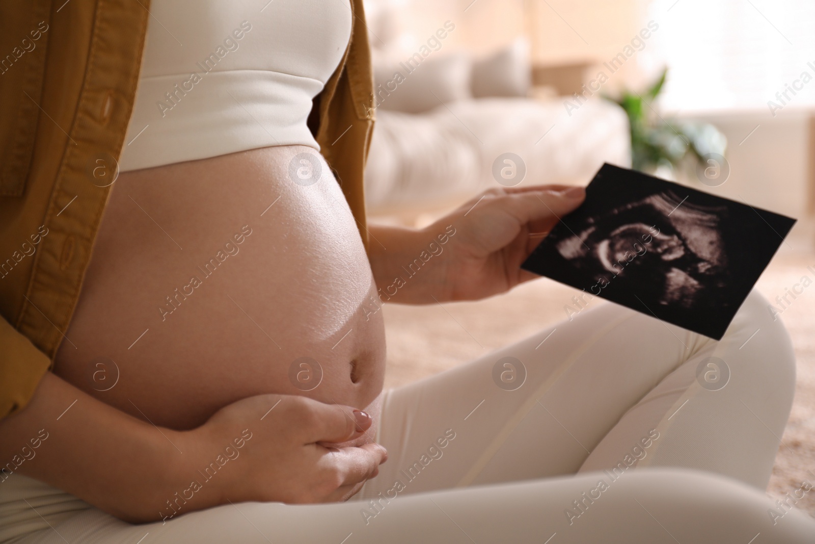 Photo of Pregnant young woman holding ultrasound picture near her belly at home, closeup