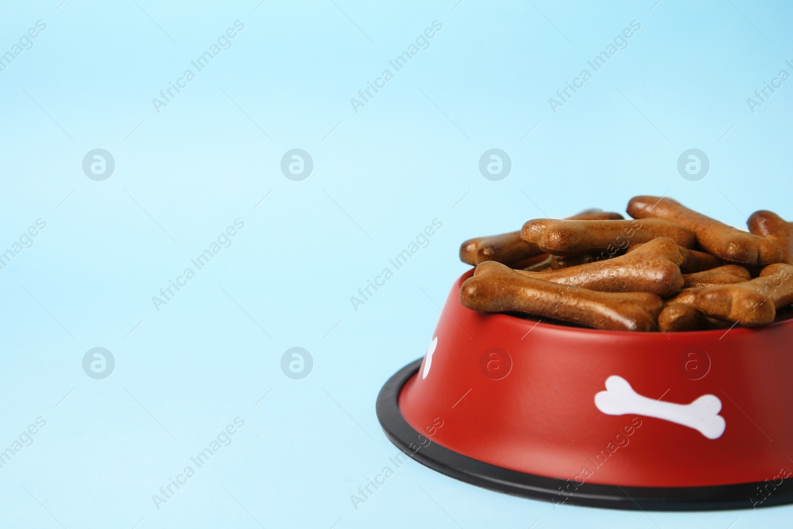 Photo of Red bowl with bone shaped dog cookies on light blue background, closeup. Space for text