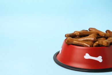 Photo of Red bowl with bone shaped dog cookies on light blue background, closeup. Space for text