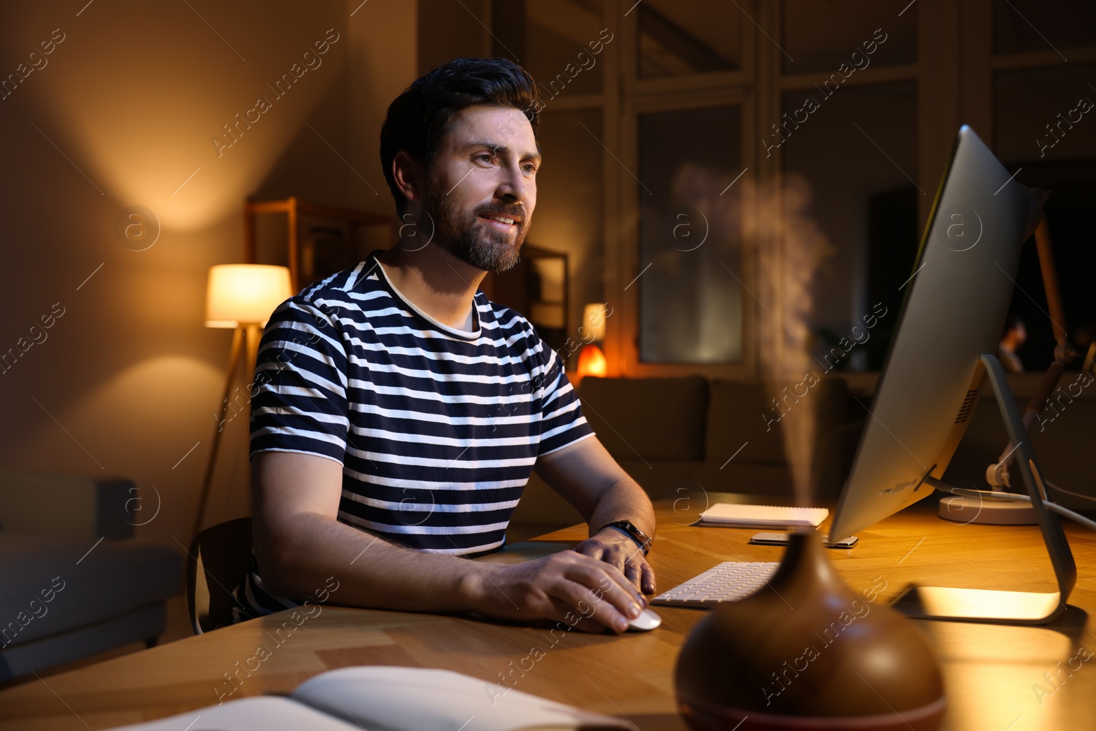 Photo of Home workplace. Man working with computer at wooden desk in room at night