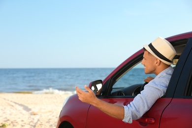 Happy man leaning out of car window on beach, space for text. Summer trip