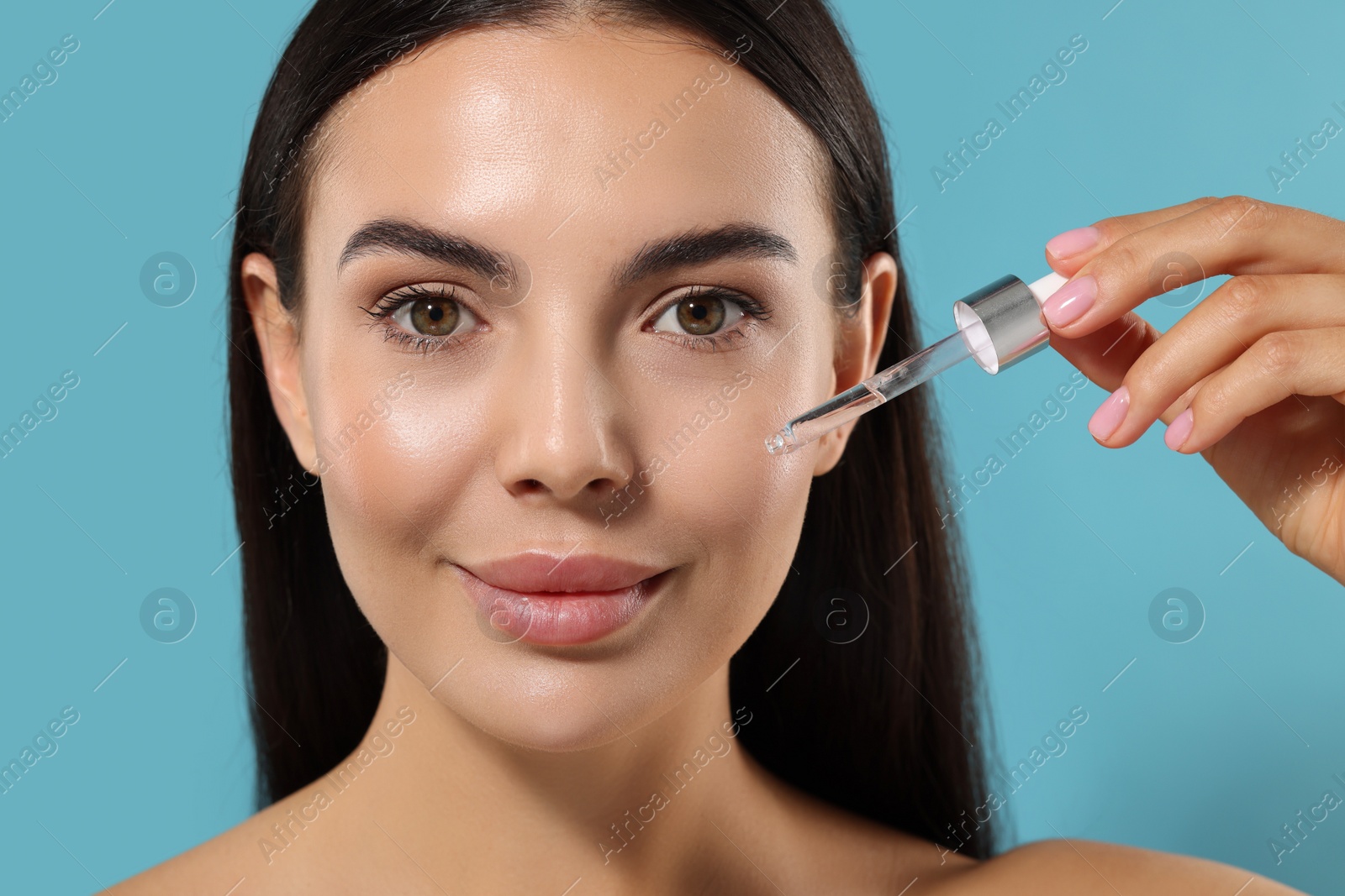 Photo of Beautiful young woman applying serum onto her face on light blue background, closeup