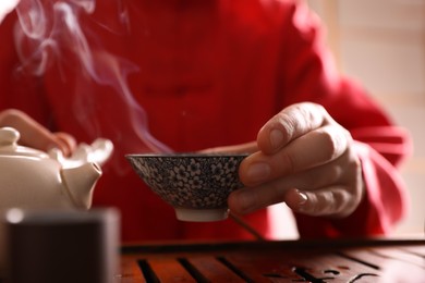 Photo of Master holding cup of freshly brewed tea during traditional ceremony at table indoors, closeup