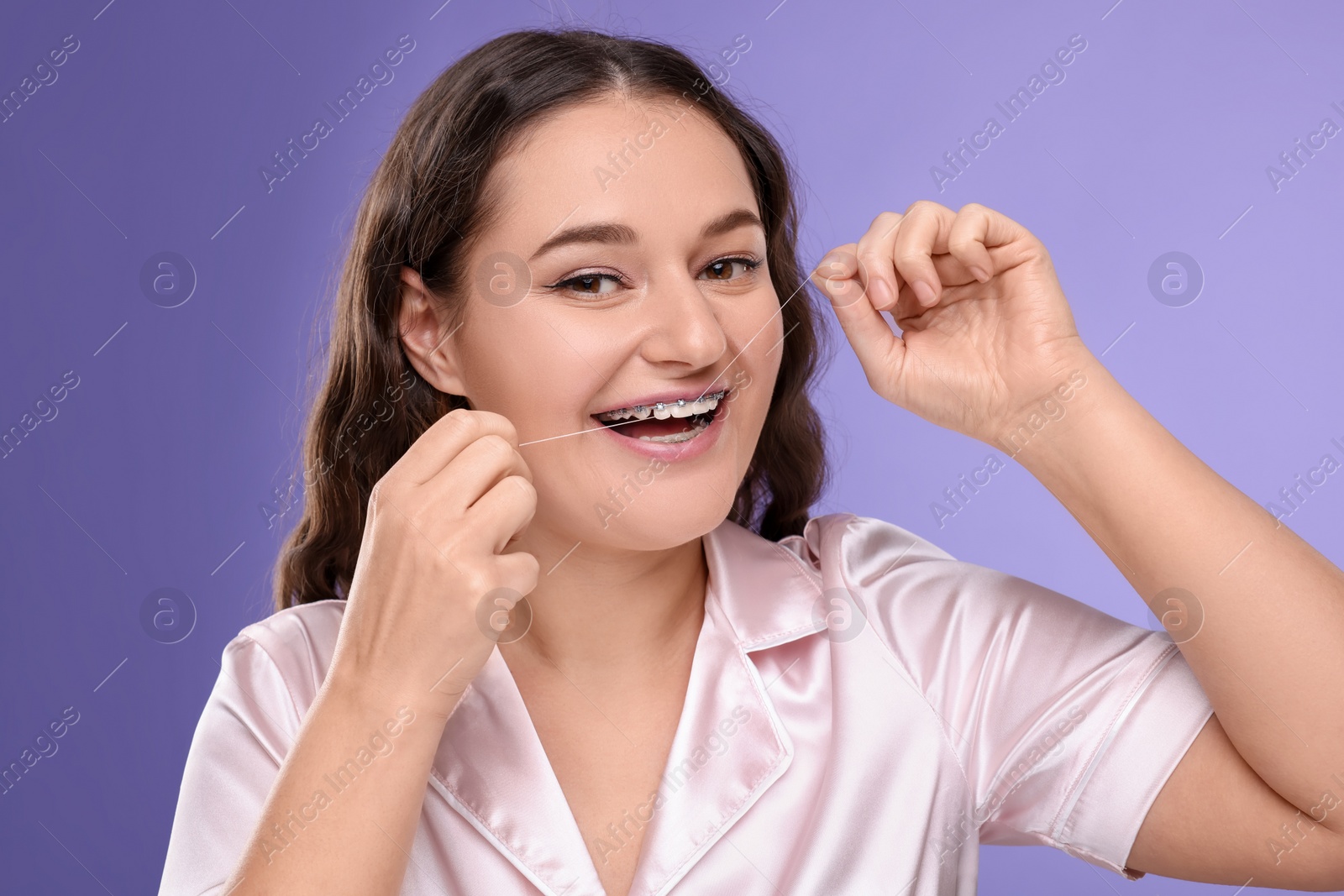 Photo of Woman with braces cleaning teeth using dental floss on violet background