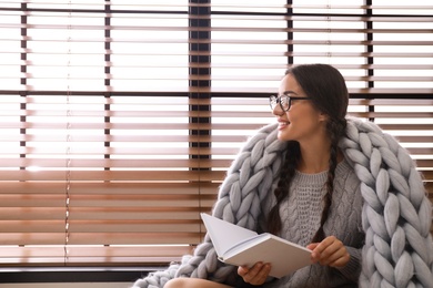 Image of Happy young woman wrapped in knitted blanket with book near window. Lazy morning