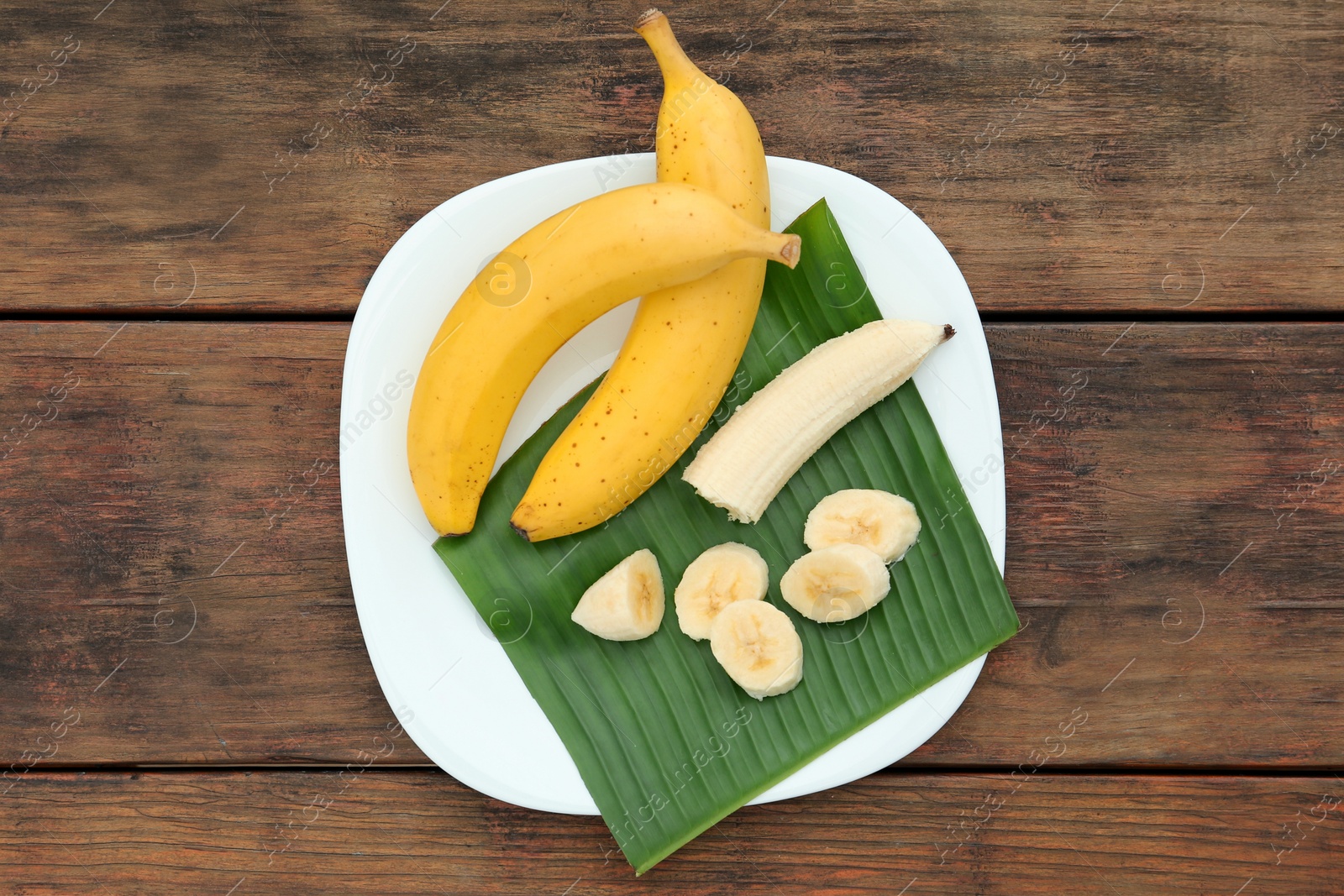 Photo of Plate with delicious bananas and fresh leaf on wooden table, top view