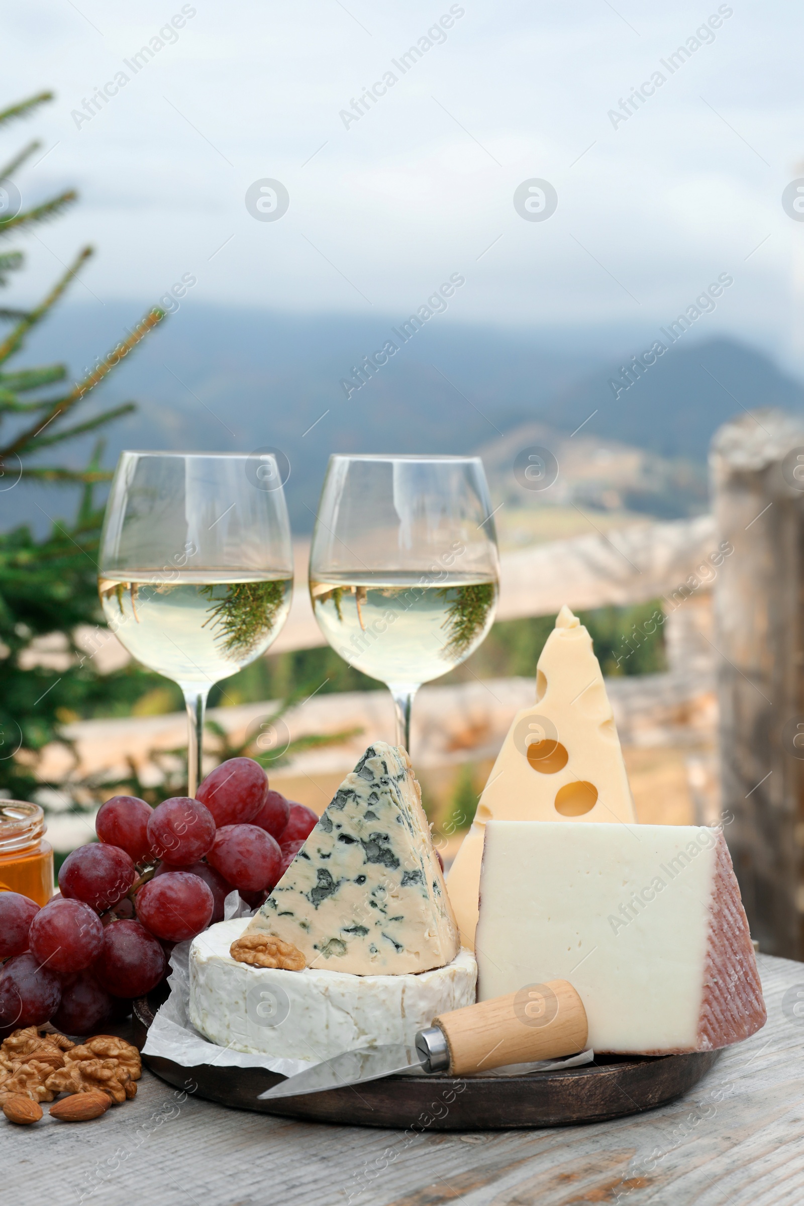 Photo of Different types of delicious cheeses, snacks and wine on wooden table against mountain landscape